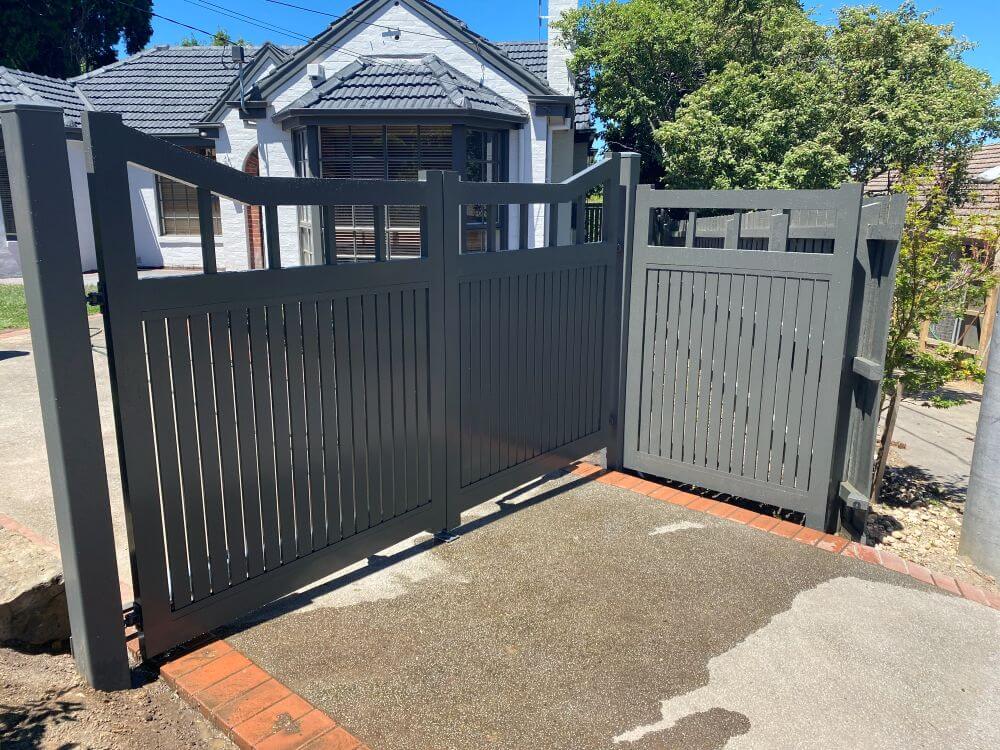 Angled street view of custom Monument aluminium driveway gates complementing the home's dark roofline and white render.